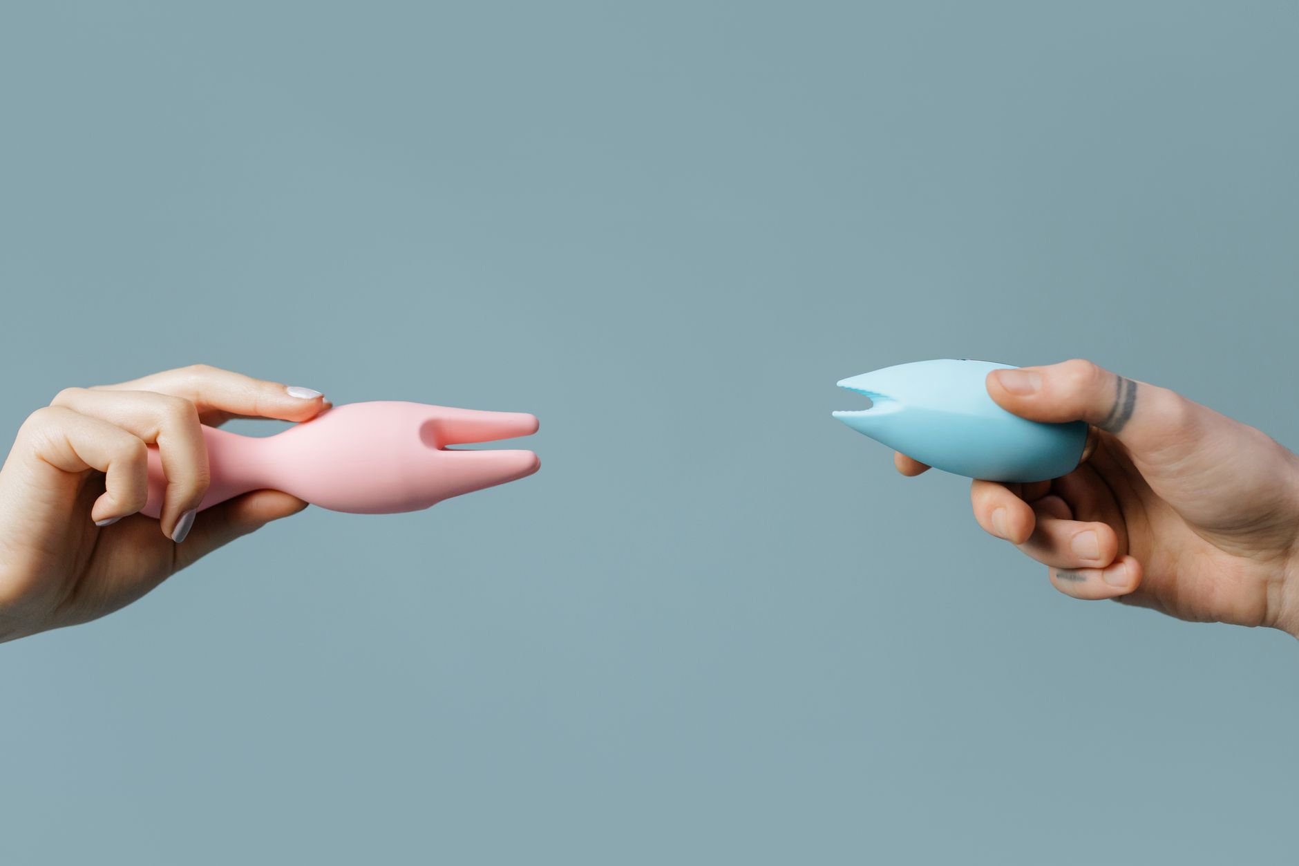 Two hands holding pink and blue clitoral vibrators against a pastel background, representing pleasure during hormonal transition.